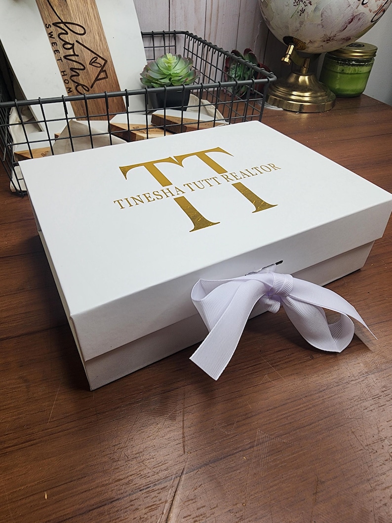 May include: A white gift box with gold lettering that reads "TT TINESHA TUTT REALTOR". The box is tied with a white ribbon. The box is on a wooden surface.