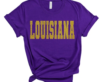 Puede incluir: Camiseta morada con la palabra "LOUISIANA" en letras doradas con purpurina.
