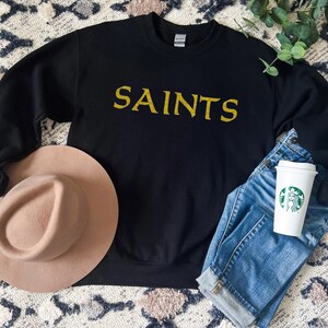 May include: Black sweatshirt with gold glitter lettering that says "SAINTS". The sweatshirt is laid flat on a patterned surface with a brown hat and blue jeans.