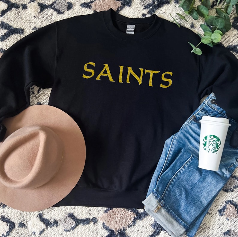 May include: Black sweatshirt with gold glitter lettering that says "SAINTS". The sweatshirt is laid flat on a patterned surface with a brown hat and blue jeans.