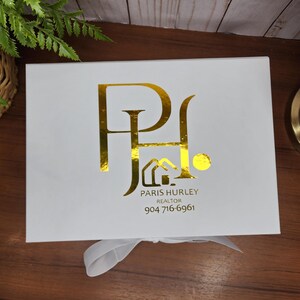 May include: A white gift box with a gold foil logo and text. The logo features the letters "P" and "H" intertwined. Below the logo is the text "PARIS HURLEY REALTOR" and a phone number.