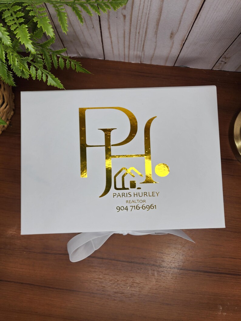 May include: A white gift box with a gold foil logo and text. The logo features the letters "P" and "H" intertwined. Below the logo is the text "PARIS HURLEY REALTOR" and a phone number.