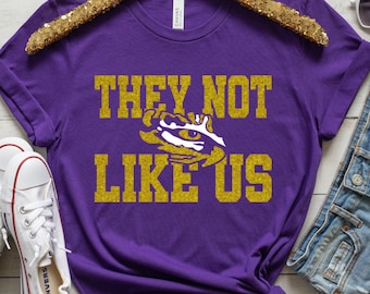 Puede incluir: Camiseta morada con texto en purpurina dorada que dice "They Not Like Us" y una huella de pata de tigre LSU en purpurina dorada.