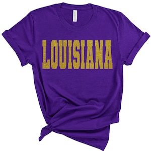 K&ouml;nnte beinhalten: Lilafarbenes T-Shirt mit dem Wort "LOUISIANA" in goldenen Glitzerbuchstaben.