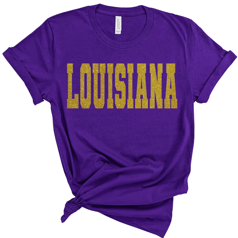 K&ouml;nnte beinhalten: Lilafarbenes T-Shirt mit dem Wort "LOUISIANA" in goldenen Glitzerbuchstaben.