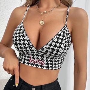 May include: A black and white houndstooth crop top with thin straps and a plunging neckline. The word "bama" is embroidered in pink across the bottom. The top is paired with black jeans and gold necklaces.