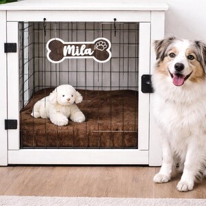 Pode incluir: Uma gaiola de madeira branca para cães com uma porta de arame preta e uma cama marrom no interior. Um brinquedo de pelúcia branco para cães está sobre a cama. Uma placa de madeira em forma de osso com o nome "Mila" está pendurada na porta. Um cão está sentado ao lado da gaiola.