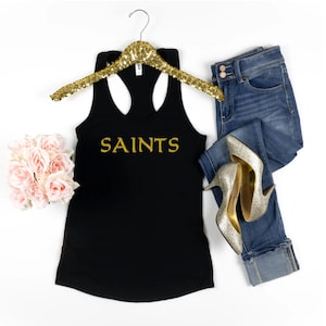 May include: Black tank top with gold glitter lettering that says "SAINTS". The tank top is on a hanger and there are jeans and gold heels in the background.