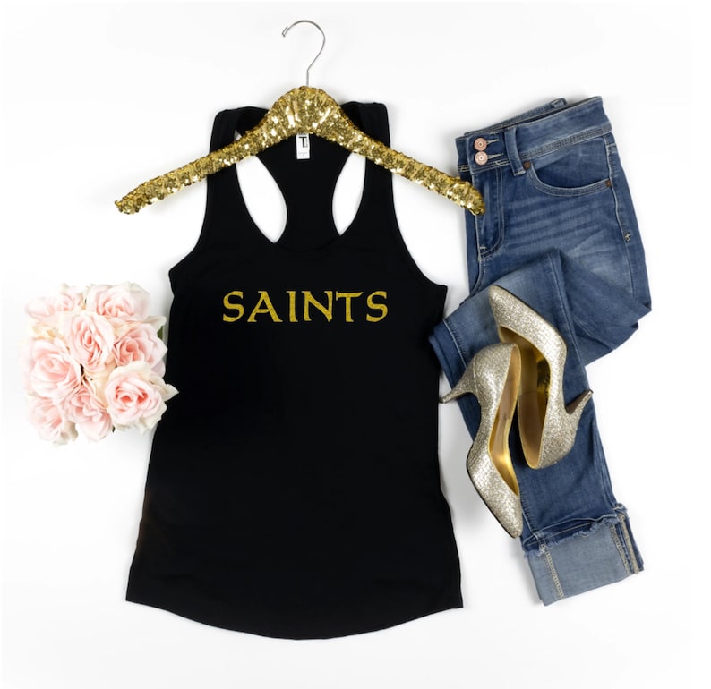 May include: Black tank top with gold glitter lettering that says "SAINTS". The tank top is on a hanger and there are jeans and gold heels in the background.