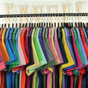 K&ouml;nnte beinhalten: Ein Gestell mit T-Shirts in verschiedenen Farben, darunter Neonorange, Neongr&uuml;n, Neongelb, S&auml;urelila, Korallen Seide, Texas Orange, Saphir, Neonblau, Irisch Gr&uuml;n, Orange, Beere, Brombeere, Kobalt, Azalee, Seidenmais, Heide-Orchidee, Mitternachtsblau, G&auml;nsebl&uuml;mchen, Violett, K&ouml;nigsblau, Schwarz, Heliconia, Rasen Gr&uuml;n, Carolina Blau, Rot, Indigo Blau, Lila, Tropisches Blau, Hellblau, Himmelblau, Maroon und Sport Grau.