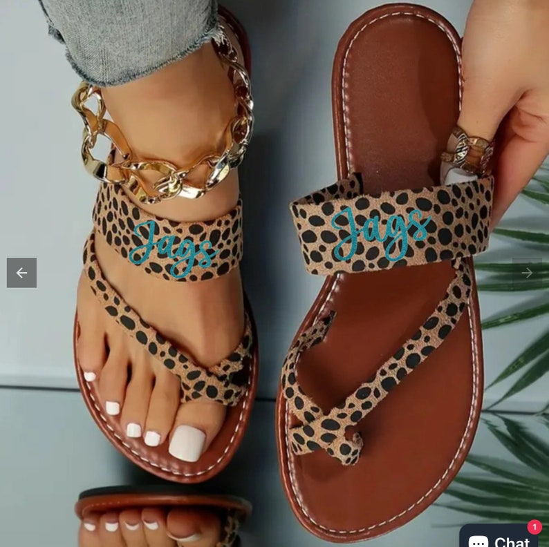 May include: A pair of brown leather sandals with a leopard print design. The sandals have a thong strap and a single strap across the top of the foot. The strap has the word "JAGS" embroidered in turquoise.