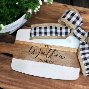May include: A white and brown wooden chopping board with a black and white checked ribbon tied around it. The board is engraved with "The Walker Family".