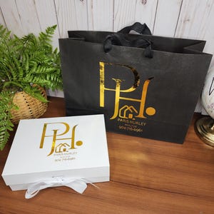 May include: A white gift box with a white ribbon and a black shopping bag. Both items feature a gold logo with the text "Paris Hurley Realtor" and a phone number. The items are on a wooden surface.