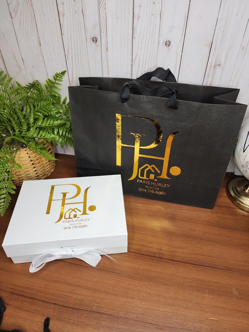 May include: A white gift box with a white ribbon and a black shopping bag. Both items feature a gold logo with the text "Paris Hurley Realtor" and a phone number. The items are on a wooden surface.