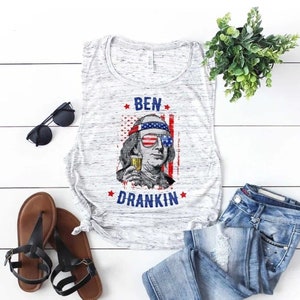 May include: A white tank top with a distressed look featuring a graphic of Benjamin Franklin wearing sunglasses and holding a beer. The graphic is surrounded by stars and the text "BEN DRANKIN".