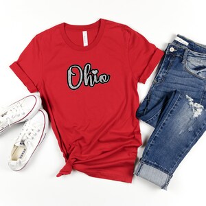 Könnte beinhalten: Rotes T-Shirt mit einer glitzernden silbernen Grafik, auf der "Ohio" mit einem kleinen roten Herzen steht. Das Shirt wird mit einer blauen Jeans und weißen Turnschuhen kombiniert.