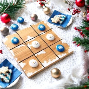 Könnte beinhalten: Ein hölzernes Tic-Tac-Toe-Spielbrett mit weiß bemalten Schneemännern und blauen Schneeflocken. Das Brett ist von Weihnachtsdekorationen umgeben, darunter Ornamente, ein kleiner Weihnachtsbaum und eine blau-weiß gemusterte Serviette.