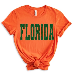 K&ouml;nnte beinhalten: Orangefarbenes T-Shirt mit dem Wort "FLORIDA" in gr&uuml;nem Glitzer-Schriftzug.