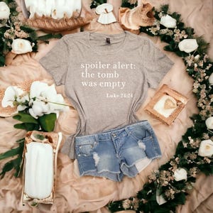 Pode incluir: Camiseta cinza com o texto "spoiler alert: the tomb was empty Luke 24:24" impresso nela. A camiseta está sobre uma superfície marrom claro com flores brancas e um short jeans azul.