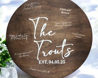 May include: A round wooden sign with the text "The Trouts EST. 04.05.25" in white lettering. The sign is decorated with various messages written in white, including "Happy Wedding Day! The Morris Family", "Congratulations! Malaca & Travis", "I love you! Jessica", "Have a great life! Marc and Karman", "To many happy years! Anthony", "To many happy years! The Martins", and "Happy ever after".