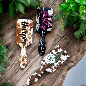 May include: Three hairbrushes with cowhide print and personalized names. The names are "Lilly", "Harper", and "G" in different colors.