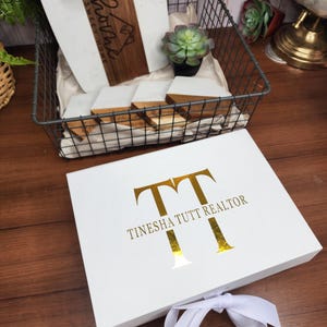 May include: A white gift box with gold lettering that reads "TT TINESHA TUTT REALTOR" and a white ribbon tied in a bow. A wire basket holds a wooden cutting board, coasters, and a small succulent.