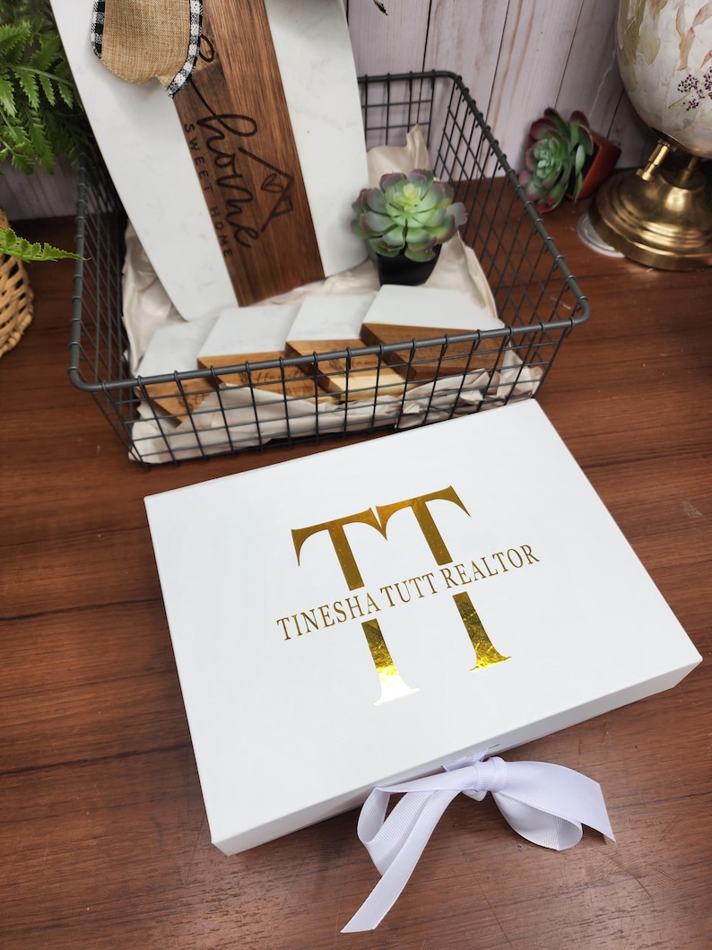 May include: A white gift box with gold lettering that reads "TT TINESHA TUTT REALTOR" and a white ribbon tied in a bow. A wire basket holds a wooden cutting board, coasters, and a small succulent.