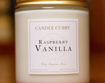 May include: A white candle with a gold lid and a label that reads "CANDLE CUBBY RASPBERRY VANILLA Bring Happiness Home  Soy  Scented Candle Made in Crystal Lake, IL"