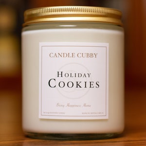 May include: A white candle jar with a gold lid and a label that reads "CANDLE CUBBY HOLIDAY COOKIES Bring Happiness Home" and "Hand-poured Scented Candle Made in Crystal Lake, IL"