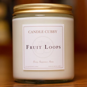 May include: A white candle in a glass jar with a gold lid. The label reads "CANDLE CUBBY FRUIT LOOPS Bring Happiness Home" and "SCENTED CANDLE MADE IN CRYSTAL LAKE, IL."