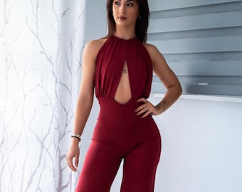 Pode incluir: Macacão borgonha com gola halter e recorte em forma de fechadura no peito. As calças de pernas largas e o corpete ajustado criam uma silhueta elegante. A modelo está em frente a uma janela com uma cortina estampada.