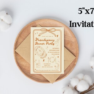Puede incluir: Una invitaci&oacute;n de 5"x7" para una cena de Acci&oacute;n de Gracias entre amigos. La invitaci&oacute;n es beige con texto e ilustraciones marrones, incluyendo un pavo, vino y cubiertos. Descansa sobre un plato de madera con un sobre marr&oacute;n.