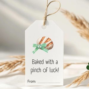 May include: A white gift tag with a watercolour illustration of a brown and white pastry with a green ribbon. The text on the tag reads "Baked with a pinch of luck!" and "From:"