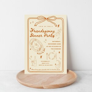 Puede incluir: Una invitaci&oacute;n crema para una fiesta de Friendsgiving con texto e ilustraciones marrones. La invitaci&oacute;n incluye la fecha, la hora y el lugar. La invitaci&oacute;n est&aacute; sobre una bandeja de madera.