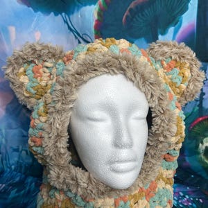 May include: A handmade, hooded scarf in a neutral color palette. The hood features plush, faux fur trim and ear-like accents. The scarf is made of a chunky, textured yarn in shades of beige, teal, and peach.