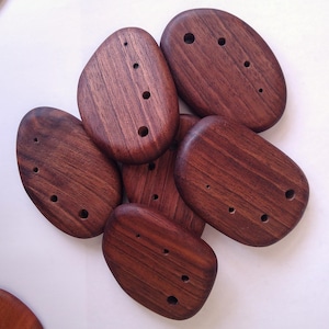 Puede incluir: Un conjunto de piedras de madera lisas de color marrón oscuro, cada una con una forma única y varios agujeros pequeños y espaciados uniformemente. La veta de la madera es visible, agregando textura. Estas piedras decorativas podrían usarse para manualidades o decoración del hogar. Medidas en centímetros.