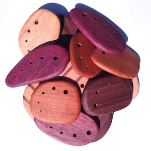 Peut inclure: Une collection de boutons en bois lisses en forme de galets, déclinés dans des tons de beige clair, brun rougeâtre, brun foncé et violet profond. Chaque bouton présente une série de petits trous percés disposés en arc de cercle.