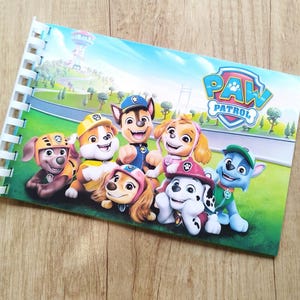 Puede incluir: Un cuaderno con espiral con los personajes de Paw Patrol. La portada muestra el logotipo de Paw Patrol e imágenes de los perros de dibujos animados sobre un campo verde con un fondo de cielo azul. El cuaderno está sobre una superficie de madera.