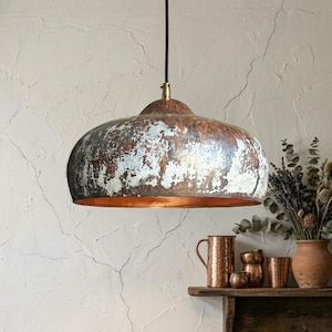 May include: A rustic, dome-shaped pendant light with a distressed, metallic finish. The light fixture has a copper-colored interior and is suspended by a black cord. The exterior shows shades of brown, silver, and white, creating a vintage aesthetic.