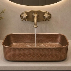 Peut inclure: Un lavabo rectangulaire en cuivre avec une texture martelée et des coins arrondis. Un robinet mural au fini laiton antique est centré au-dessus de la vasque, avec deux poignées en croix de chaque côté, distribuant un filet d'eau constant.