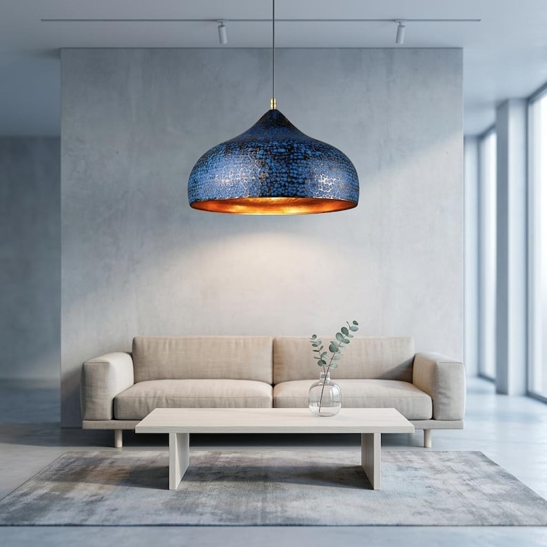 May include: A modern, blue pendant light with a textured surface hangs above a beige sofa and light wood coffee table. The interior of the light fixture is gold, and the room has a minimalist design with a neutral color palette.
