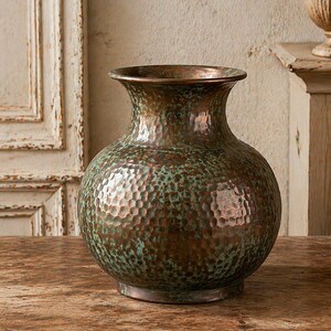 Könnte beinhalten: Eine gehämmerten Kupfervase mit bauchigem Körper und ausgestelltem Rand. Die Vase hat eine strukturierte Oberfläche mit einer gesprenkelten grün-braunen Patina. Die Vase steht auf einem runden Sockel und ist ein dekoratives Wohnaccessoire.