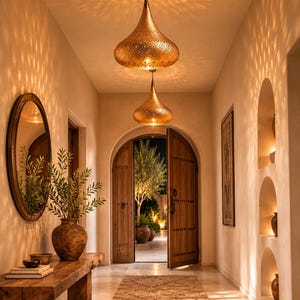 May include: Two teardrop-shaped brass pendant lights with intricate perforated patterns hang from the ceiling, casting geometric shadows on the walls. The lights illuminate a hallway featuring a wooden console table, a large vase, and a round mirror.