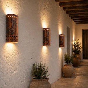 May include: Three cylindrical wall sconces with a weathered copper finish and square cutouts at the top and bottom edges. These fixtures are mounted on a textured white wall, emitting warm light upward and downward to illuminate the corridor.