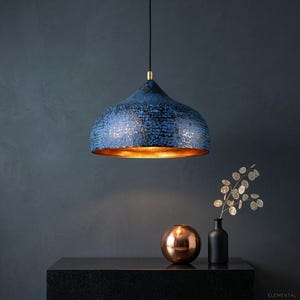 May include: A blue pendant light with a textured surface and a gold interior hangs from a black cord. The light fixture is the focal point, with a dark background and a copper-colored sphere and black vase on a black surface.