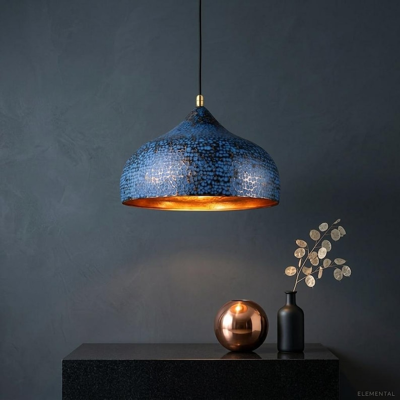 May include: A blue pendant light with a textured surface and a gold interior hangs from a black cord. The light fixture is the focal point, with a dark background and a copper-colored sphere and black vase on a black surface.