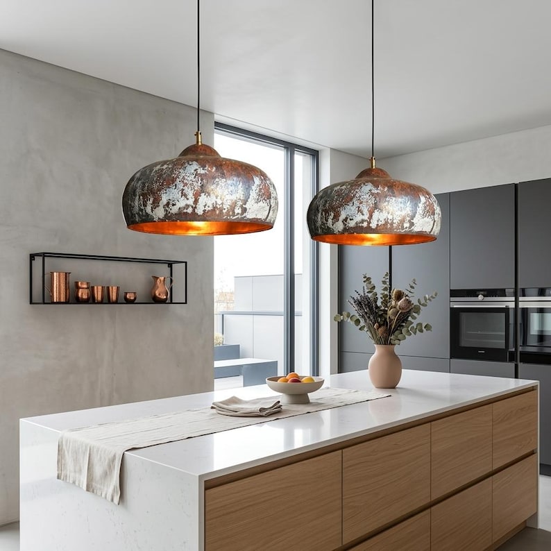 May include: Two pendant lights with distressed metal shades hang above a kitchen island. The shades have a copper-colored interior and a weathered, multi-toned exterior. The kitchen features a white countertop, wooden cabinets, and a neutral color scheme.
