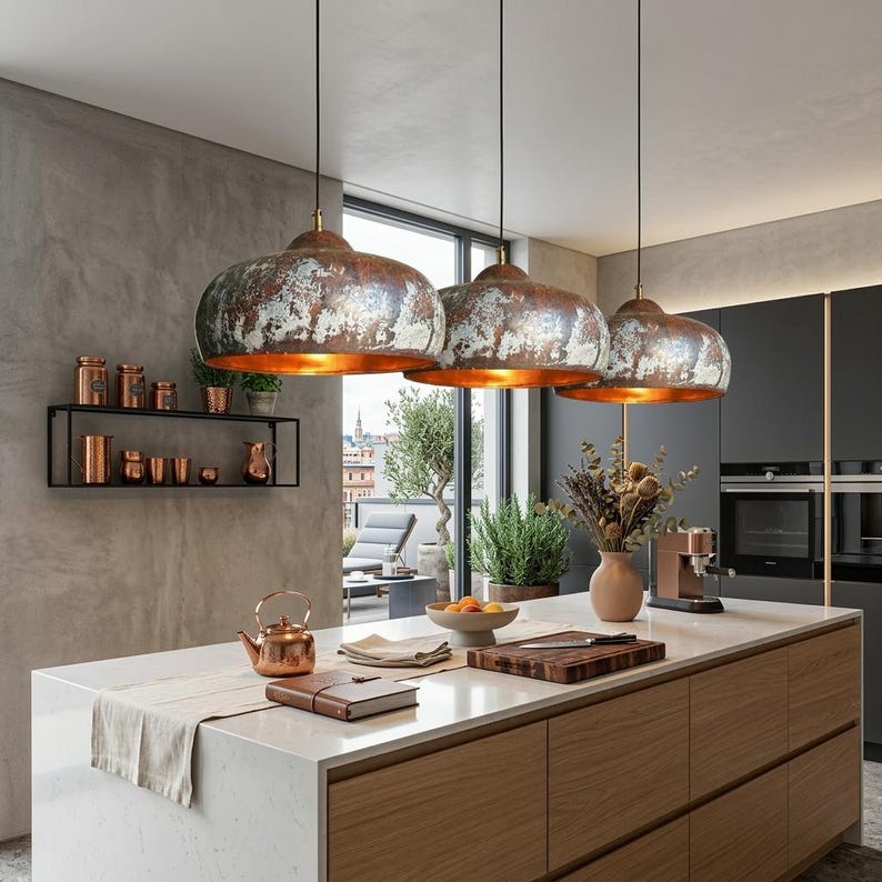 May include: Three pendant lights with distressed metal shades hang above a kitchen island. The shades have a copper interior and a weathered, mottled exterior. The kitchen features a light countertop, wooden cabinets, and a black metal shelf with copper-colored items.