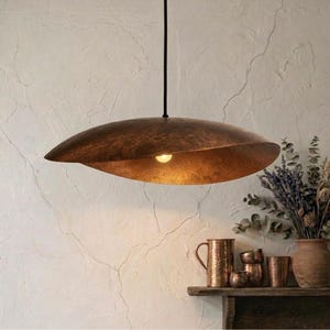 May include: A copper-colored, leaf-shaped pendant light fixture hangs from a black cord. The light fixture has a textured surface and a warm, inviting glow. The background features a white wall and a shelf with copper mugs and a vase of dried flowers.
