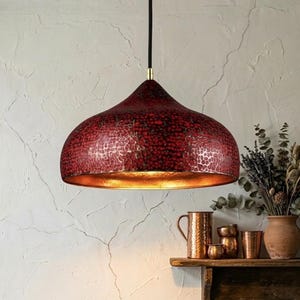 Puede incluir: Lámpara colgante roja con textura martillada e interior dorado. La lámpara, con forma de cúpula, cuelga del techo mediante un cable negro. Emite una luz cálida y acogedora. Diámetro aproximado de 40 cm.